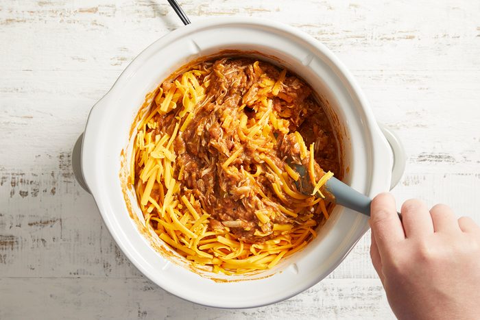 A hand stirs shredded cheese and brown seasoned meat in a white slow cooker on a light wooden surface.