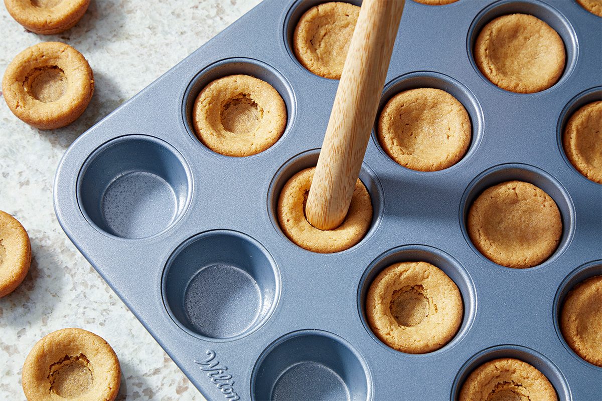 A wooden tool presses the center of a baked cookie in a muffin tin, creating an indentation; several indented cookies are in the pan and on the counter nearby.