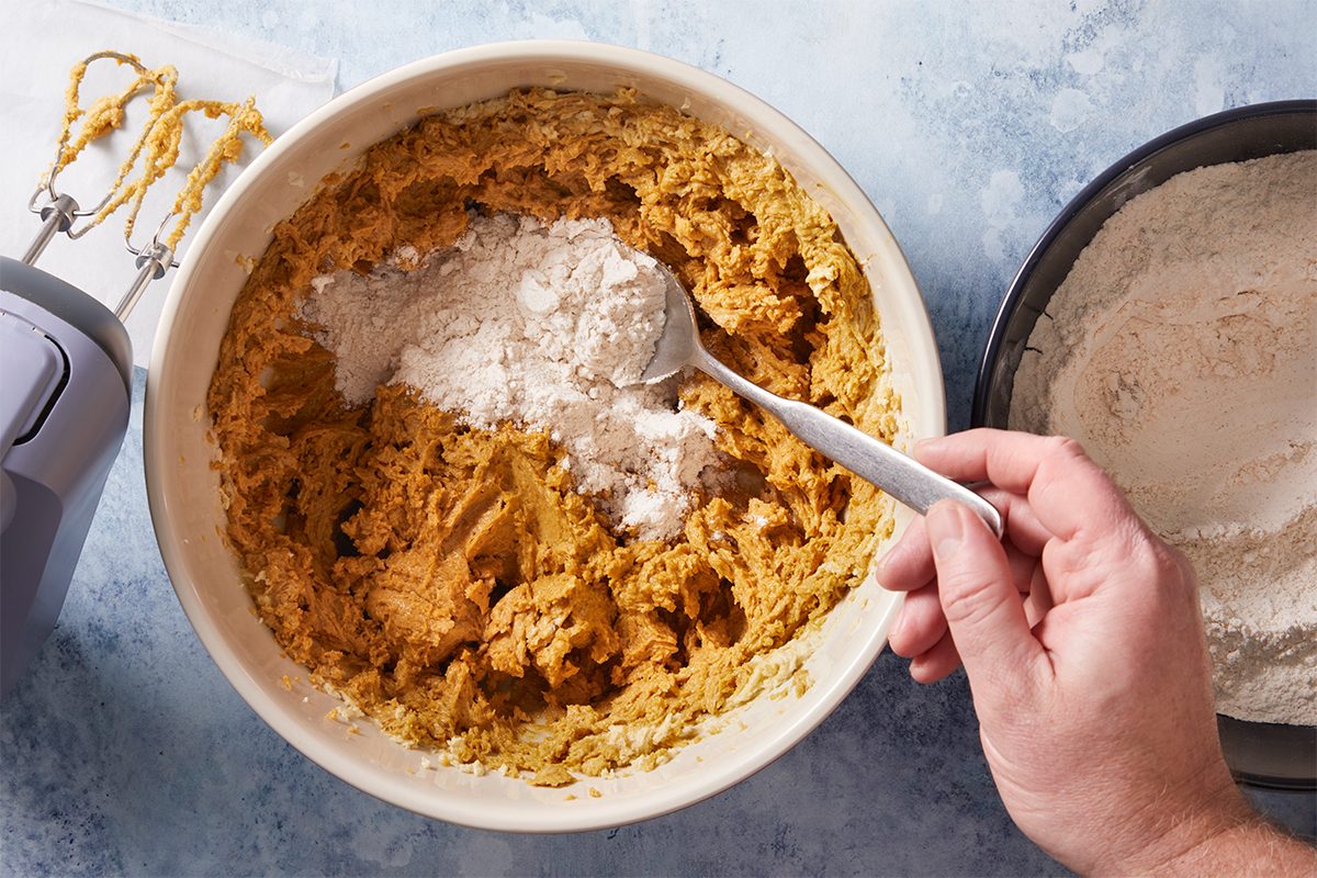 Overhead shot of a hand scoops flour into a large bowl of batter with a spoon beside it sits a bowl of flour and an electric hand mixer on blue countertop