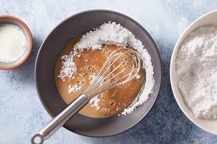 Overhead shot of a whisk is in a bowl with powdered sugar and brown liquid mixture Next to it are a cup of milk and a large bowl of powdered sugar