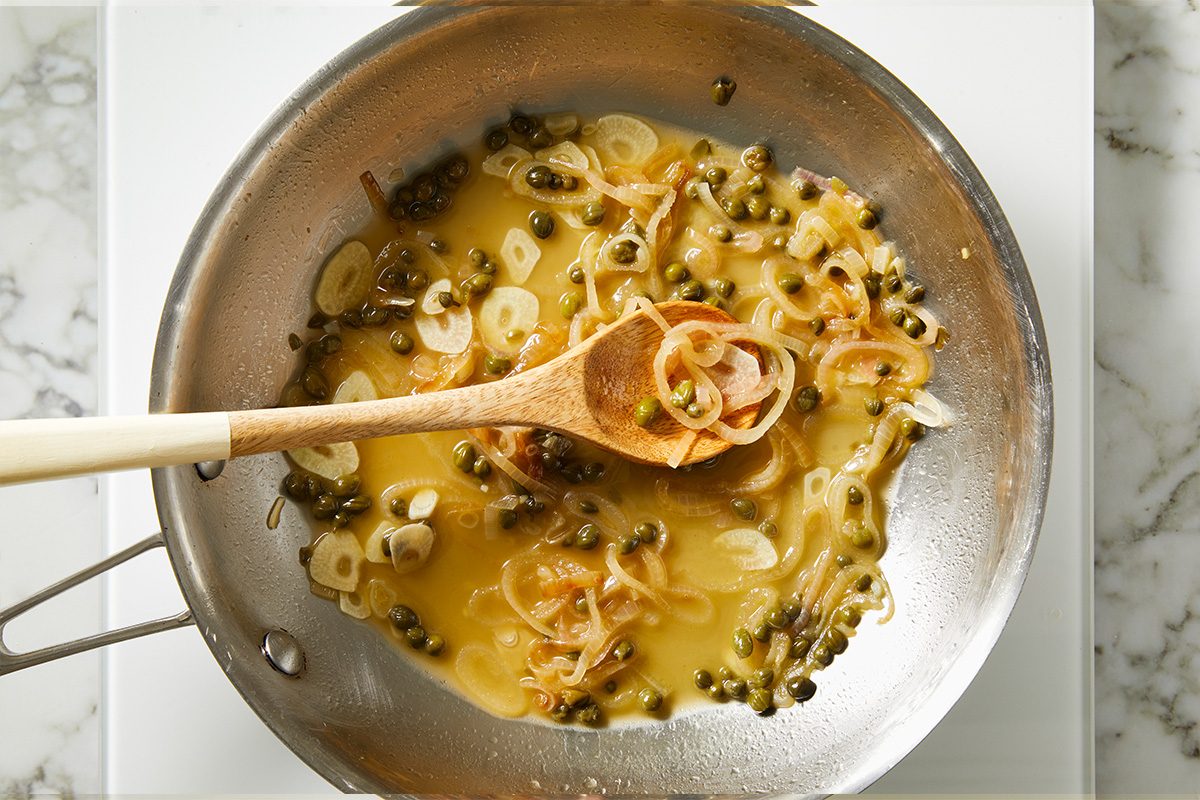 Overhead shot of a skillet with sautéed shallots, garlic, and capers simmering in a light sauce, being stirred with a wooden spoon on the stovetop;