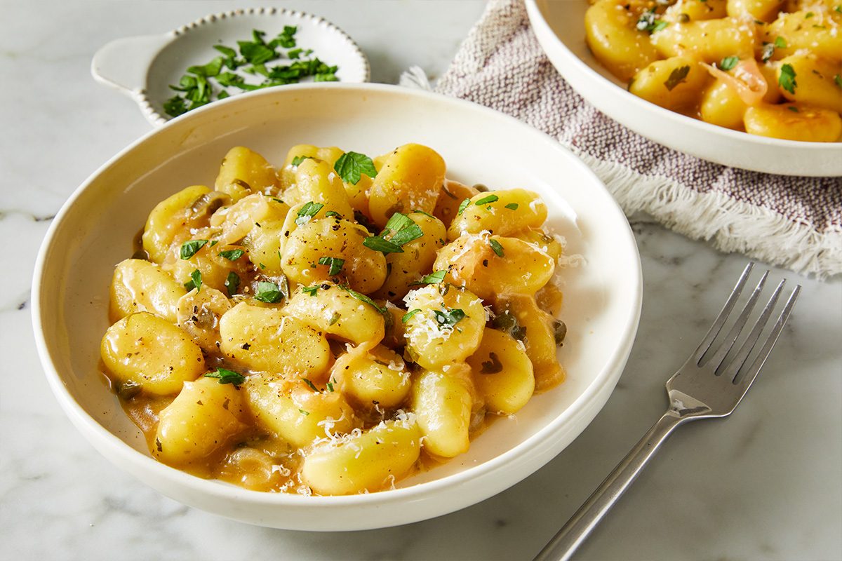 3/4 angle close-up shot of gnocchi piccata in a creamy lemon-caper sauce, garnished with fresh parsley and served in a white bowl on a light marble surface;