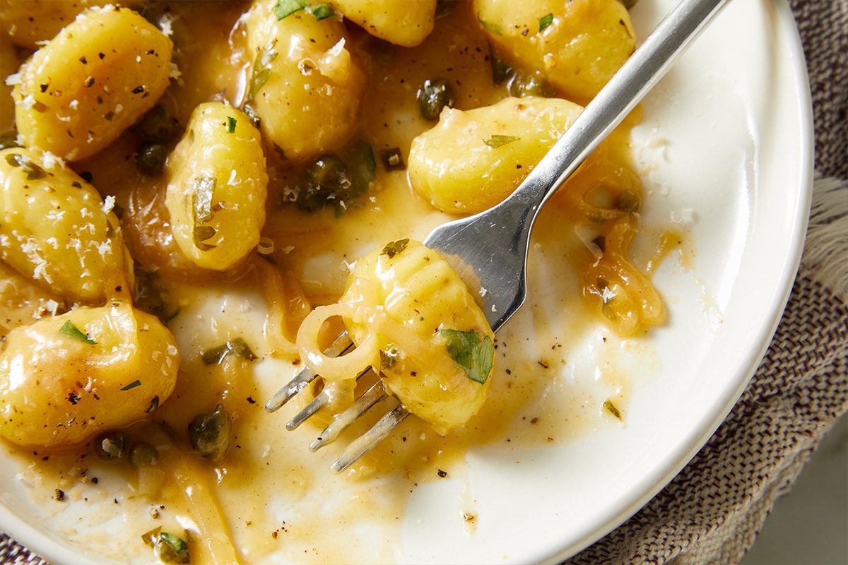 Extreme close-up shot of gnocchi piccata coated in a glossy lemon-caper sauce, with a fork lifting a tender piece from the bowl;
