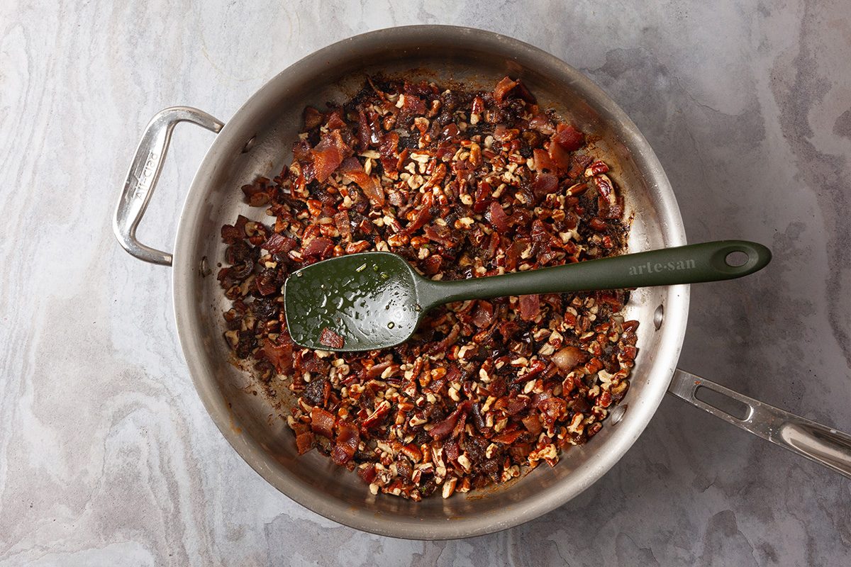 Overhead shot of a stainless steel skillet holds chopped cooked bacon and pecans on a gray marble surface A green spatula with bits rests across the pan