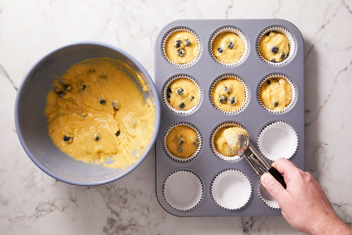 Overhead shot of a hand fills paper cupcake liners with blueberry muffin batter using a scoop and there is a mixing bowl of extra batter on the counter