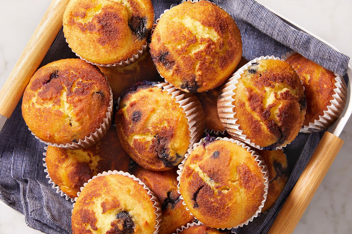 Overhead shot of a basket filled with golden-brown Keto Blueberry Muffins in paper liners and it sits on a blue cloth on a marble surface