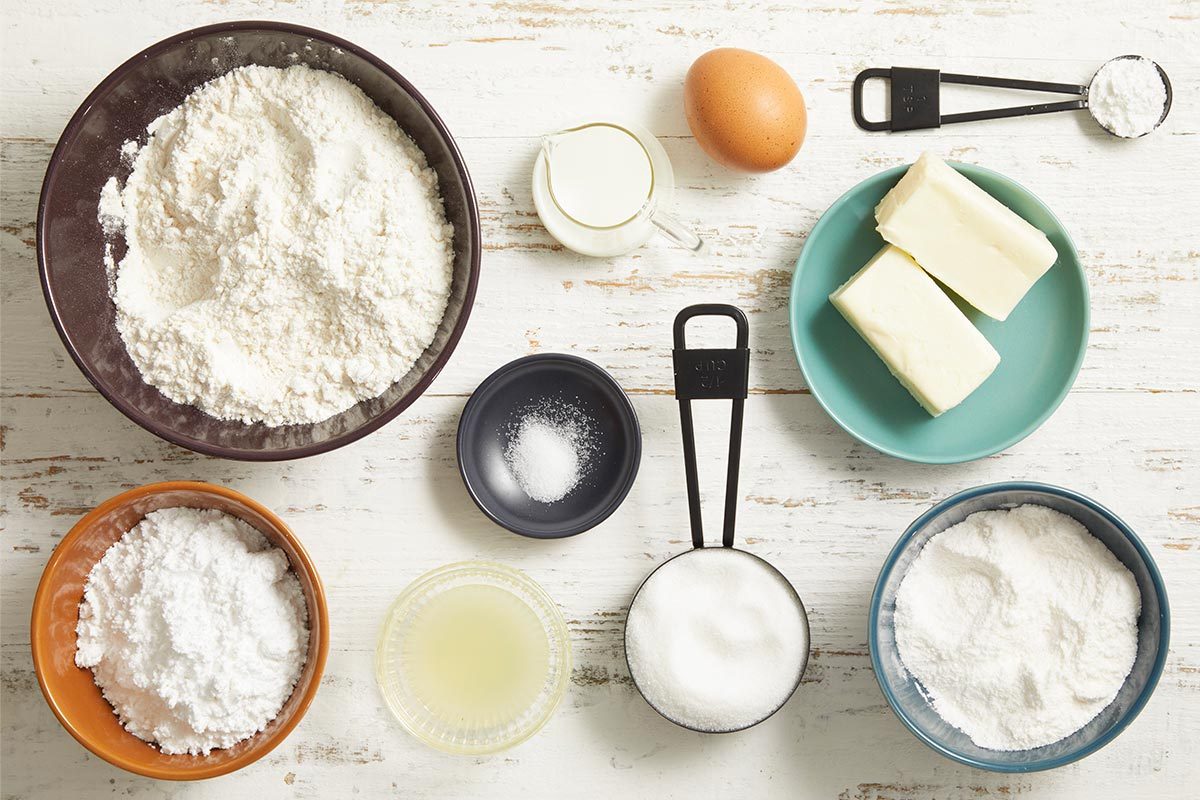 Overhead shot of cookie ingredients arranged neatly on a light surface including flour, sugar, butter, lemon pudding mix, egg, milk, and measuring spoons.