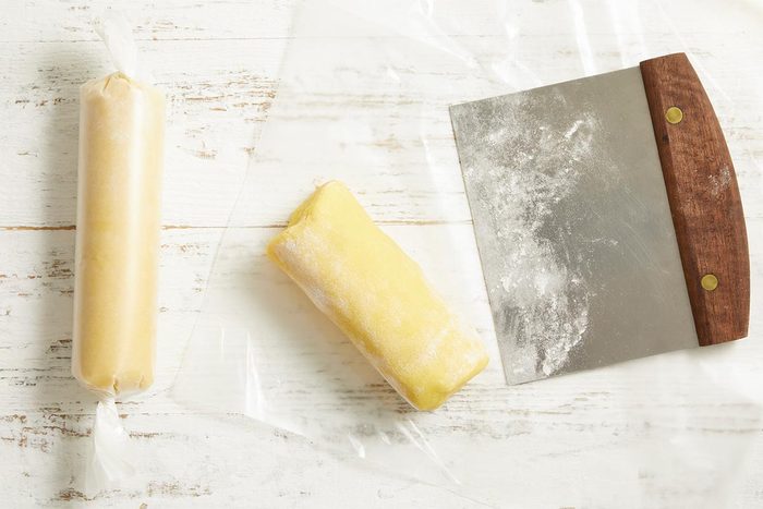 Overhead shot of rolled cookie dough logs wrapped in plastic with a bench scraper and flour scattered nearby.
