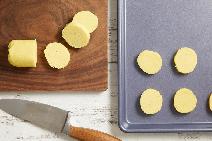 Overhead view of sliced cookie dough rounds arranged on a baking sheet, with a knife and cutting board beside it.