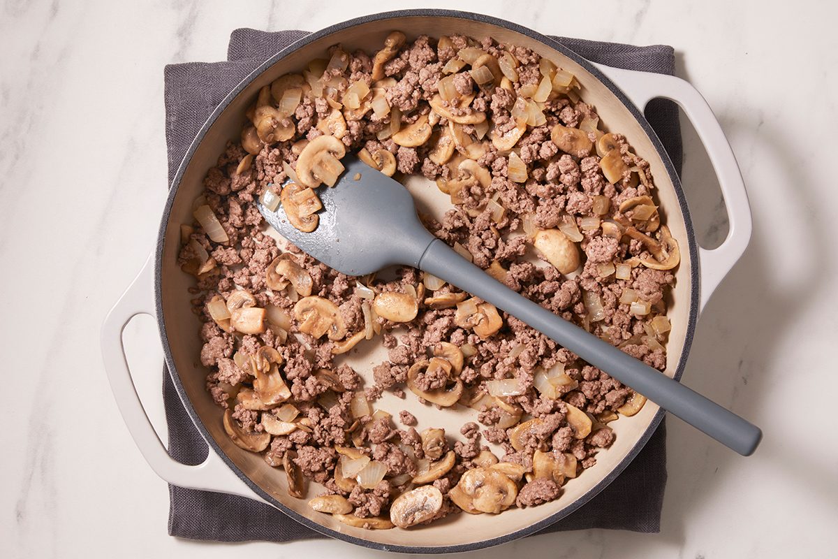 A skillet filled with cooked ground beef and sliced mushrooms sits on a dark cloth, with a gray spatula resting inside the pan.
