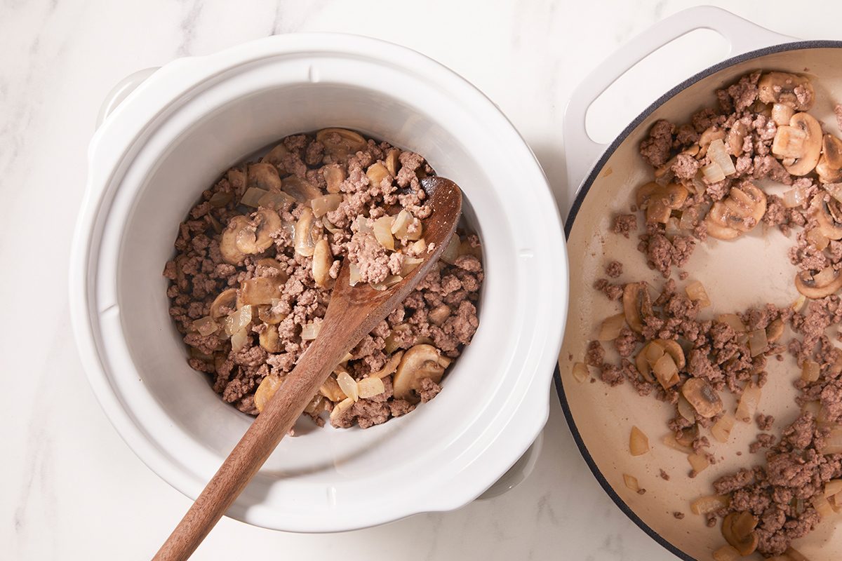 Ground meat and sliced mushrooms being stirred in a white slow cooker with a wooden spoon, next to a pot with more cooked meat and mushrooms, all on a white countertop.