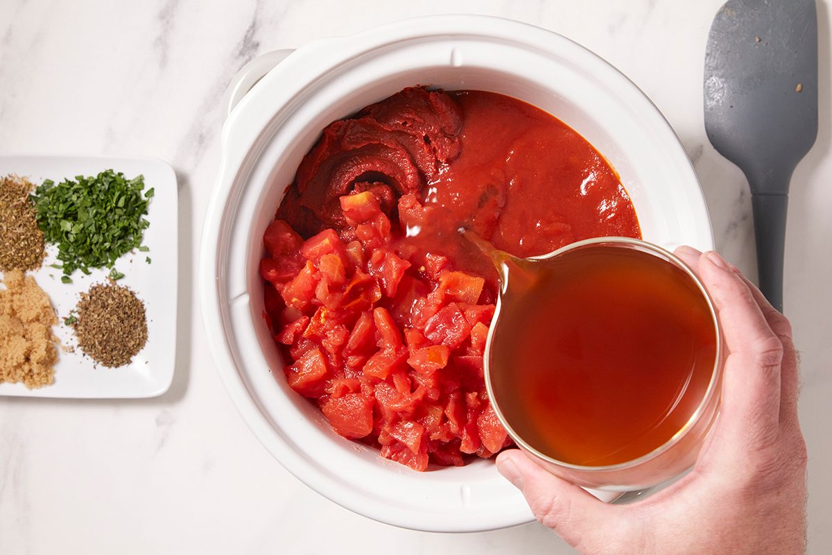 A hand pours broth into a slow cooker containing tomato sauce, diced tomatoes, and tomato paste. Nearby, a plate with measured spices and herbs sits on a white countertop.