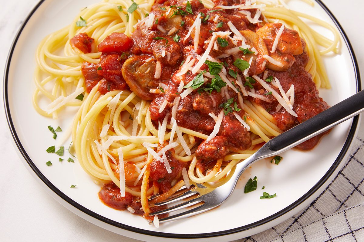 A plate of spaghetti topped with tomato sauce, sliced mushrooms, grated cheese, and chopped parsley. A fork rests on the side of the plate.