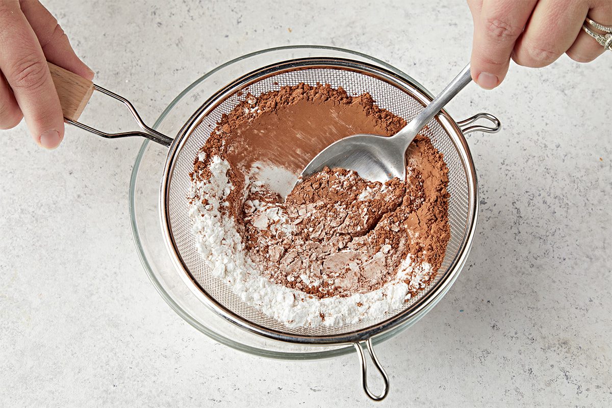 Overhead shot of a person folding whipped egg whites into a bowl of chocolate batter with a spatula on a light countertop;