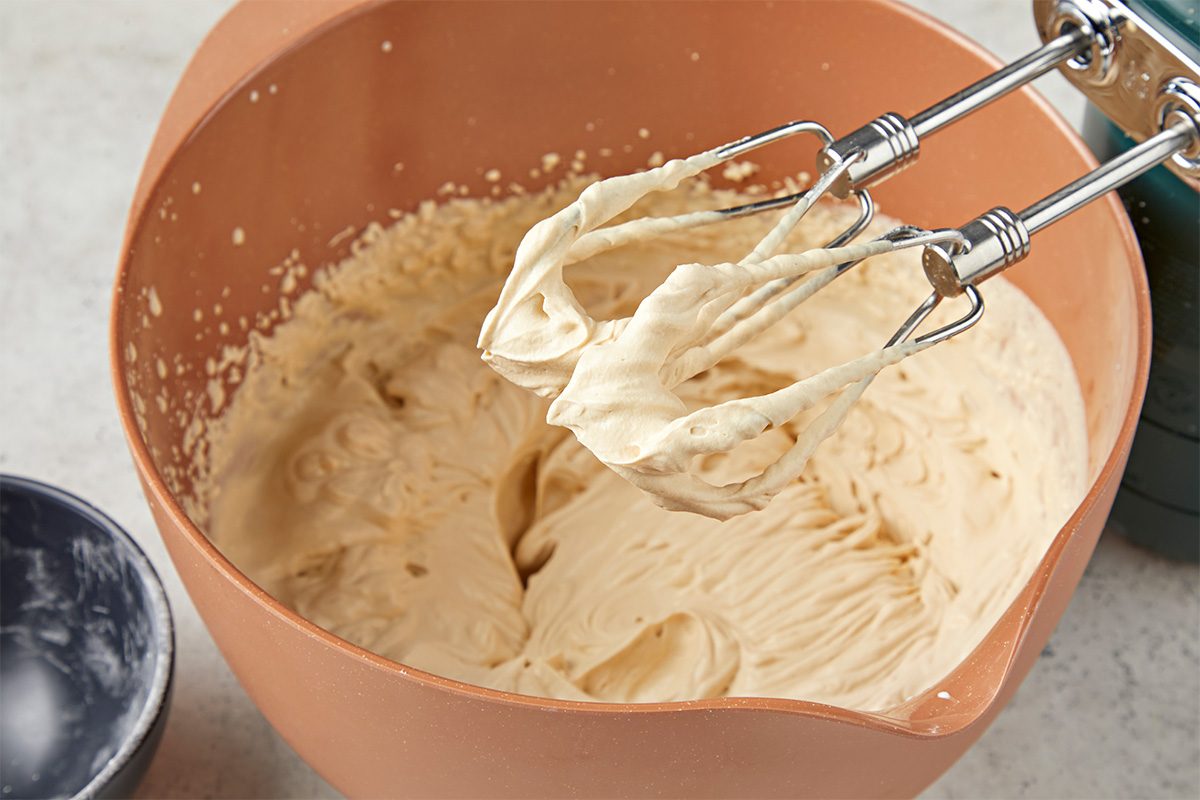 Closeup shot of an electric hand mixer with beaters covered in creamy batter, hovering over a mixing bowl filled with more batter;