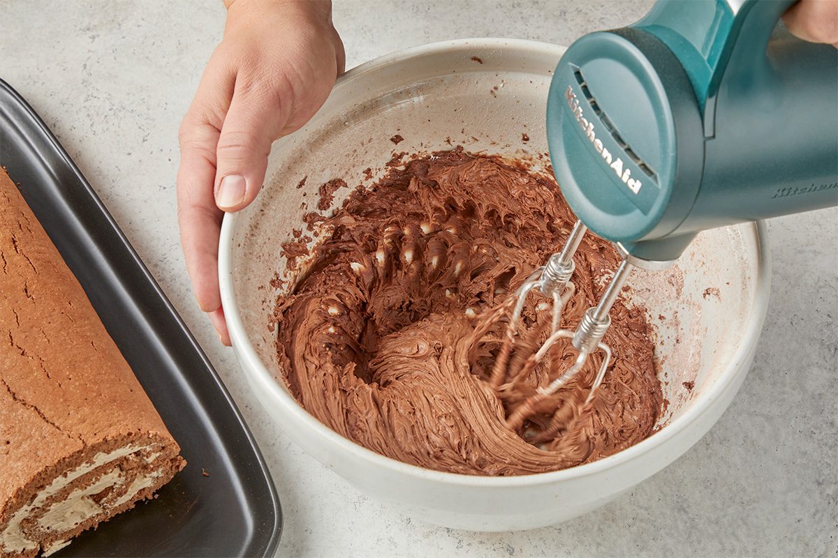 Overhead shot of a hand holding a bowl while an electric mixer blends chocolate frosting, with a partially rolled cake on a tray nearby;