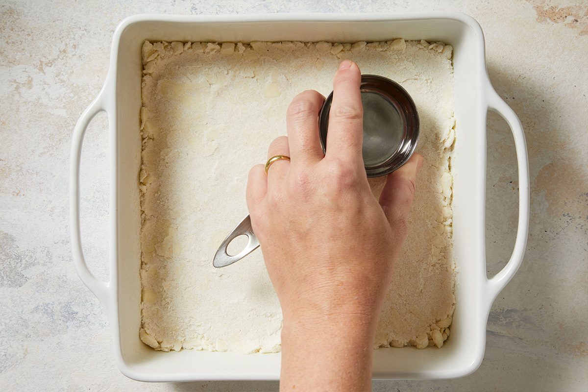Overhead view of hands pressing crust mixture into a white baking dish lined with parchment paper using a metal measuring cup.