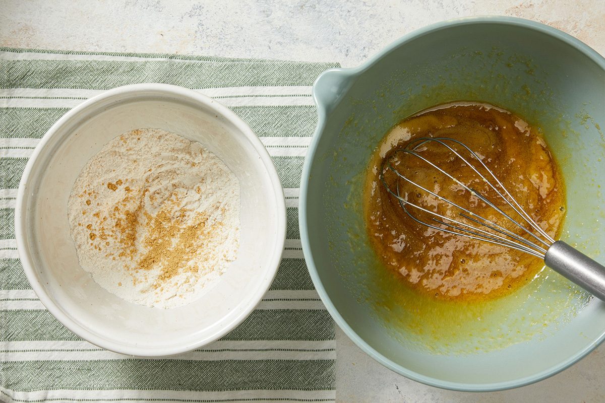 Overhead shot of wet ingredients being whisked together in a gray mixing bowl beside a smaller bowl of dry ingredients on a striped cloth.