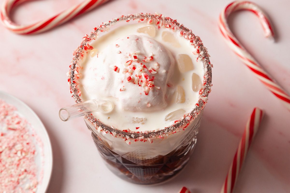 Close-up shot of a Peppermint White Russian sits in a glass topped with whipped cream and crushed peppermint with a candy cane rim and clear straw surrounded by whole and crushed candy canes on a light pink surface