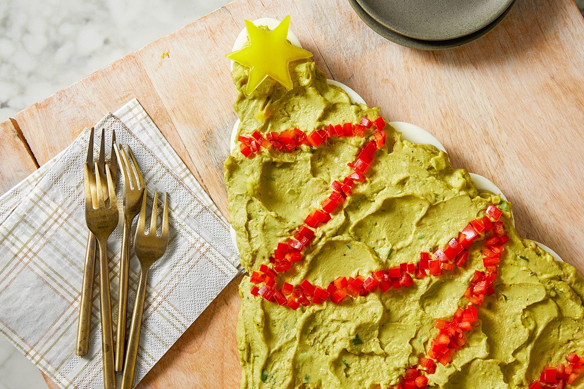 Overhead shot of a Pull-Apart Deviled Egg Christmas Tree is arranged on a Christmas tree-shaped platter topped with guacamole red pepper garland and a yellow star while three forks sit on a plaid napkin beside the wooden board
