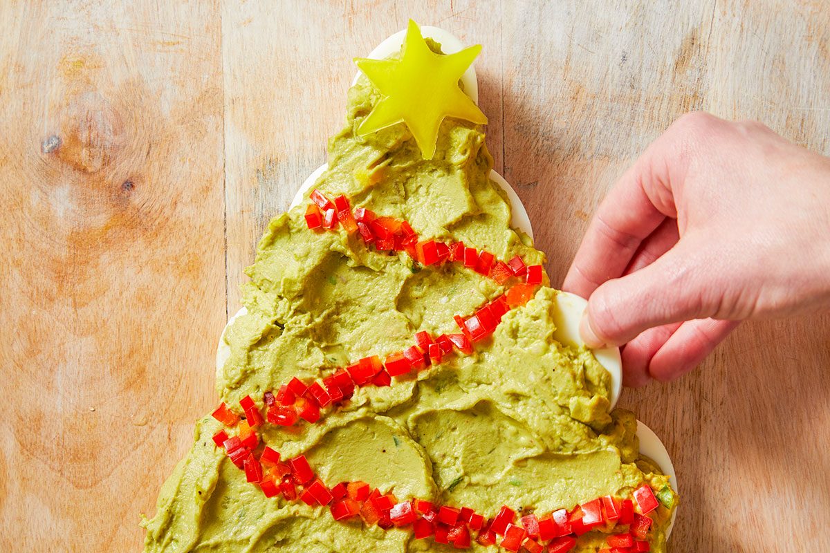 Overhead shot of a Pull-Apart Deviled Egg Christmas Tree is arranged on a Christmas tree-shaped platter topped with guacamole red pepper garland and a yellow star while three forks sit on a plaid napkin beside the wooden board
