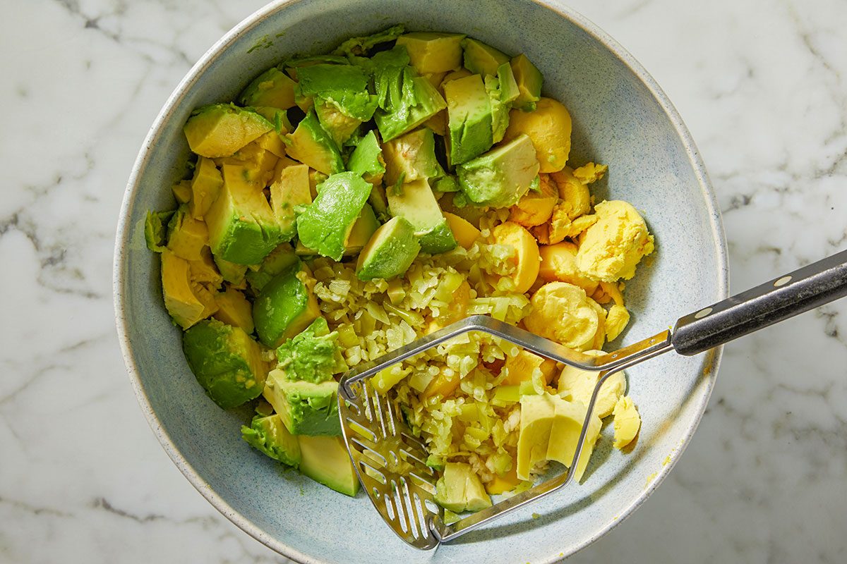 Overhead shot of a bowl holds chopped avocado and diced onions ready for mashing with a metal potato masher sitting on a white marble surface