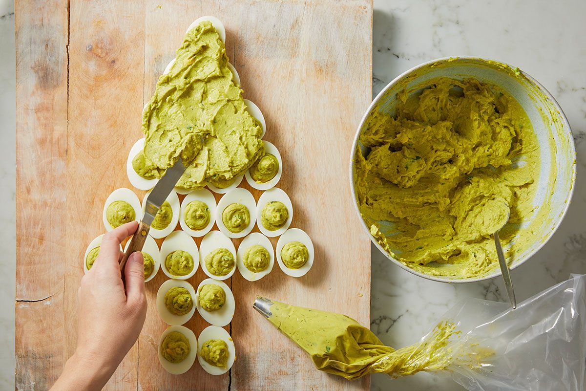 Overhead shot of a hand spreads green filling on deviled eggs arranged like a Christmas tree on a wooden board Nearby are a bowl of filling and piped bag