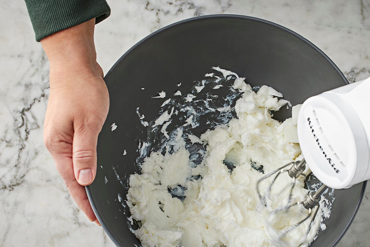 A person holds a gray mixing bowl with whipped egg whites inside while using a white electric hand mixer on a marble countertop.