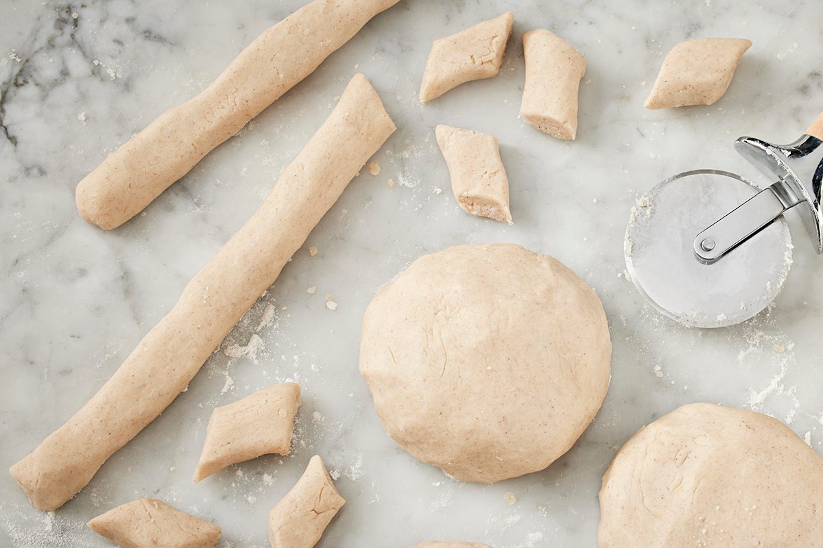 Balls and rolled logs of dough, some cut into diamond shapes, are arranged on a floured marble surface next to a pastry cutter.