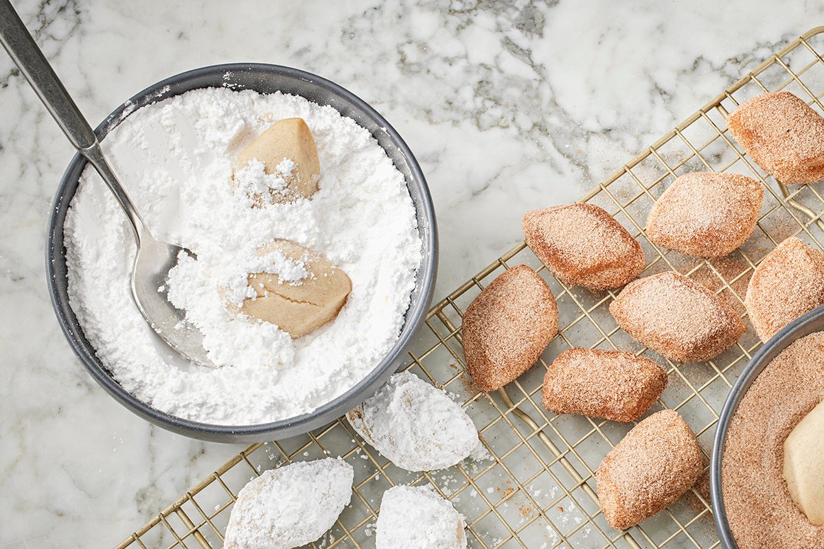 A bowl of powdered sugar with a spoon and a few small pastries being coated, next to a cooling rack with more sugar-dusted and cinnamon-coated pastries on a marble surface.