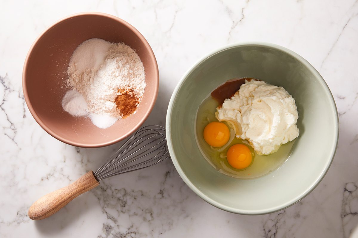 Two bowls on a marble surface: one with flour, baking powder, salt, and cinnamon; the other with two eggs and ricotta cheese. A whisk lies beside the bowls.