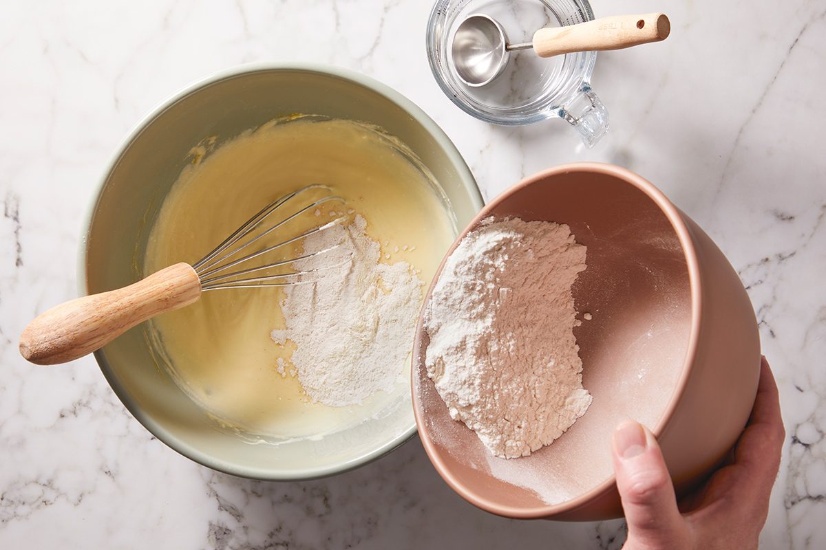 A person pours flour from a pink bowl into a larger mixing bowl with whisked batter. A whisk rests in the mixture, and a measuring cup with a spoon is nearby on a marble countertop.