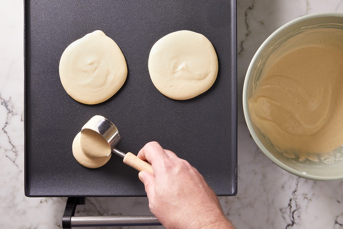 A hand pours pancake batter from a metal measuring cup onto a griddle, forming round pancakes. Two pancakes are already cooking, and a bowl of batter sits nearby on a marble countertop.