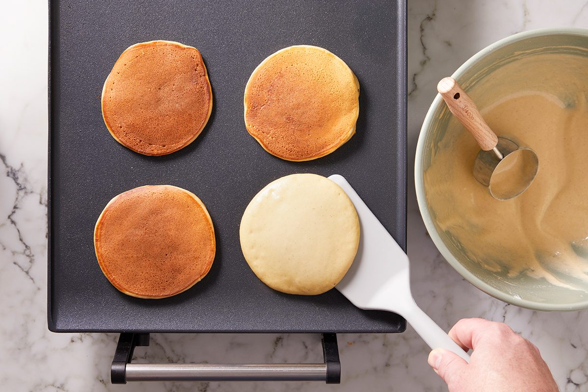Four pancakes are cooking on a griddle; three are golden brown, while one is being flipped with a spatula. A nearby bowl holds pancake batter and a wooden-handled spoon. The scene is on a marble countertop.