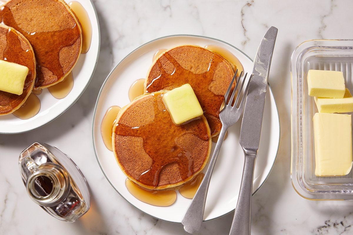 A plate with two pancakes topped with a pat of butter and syrup, next to a fork and knife. Nearby are a butter dish with slices of butter and a bottle of syrup, all on a marble surface.