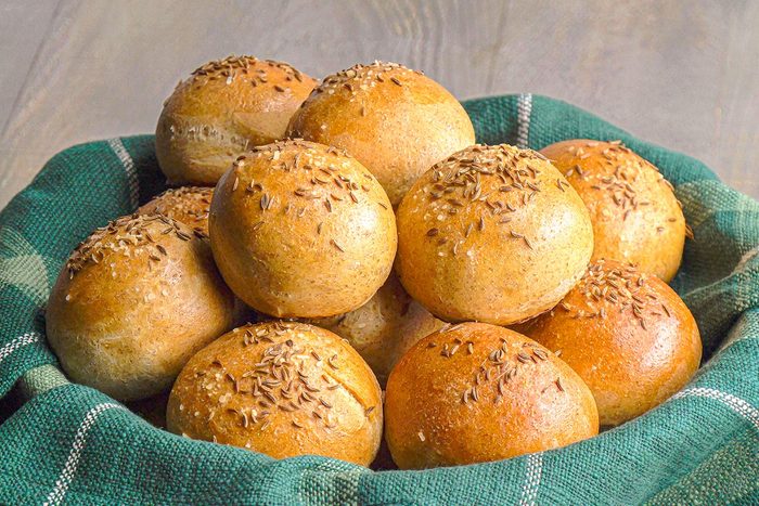 Closeup shot of a basket lined with green checkered cloth holding a pile of rye rolls round, golden brown, and topped with caraway seeds;