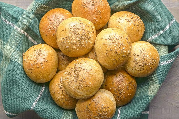 Overhead shot of a basket lined with green plaid cloth holding several rye rolls, each round and topped with caraway seeds;l