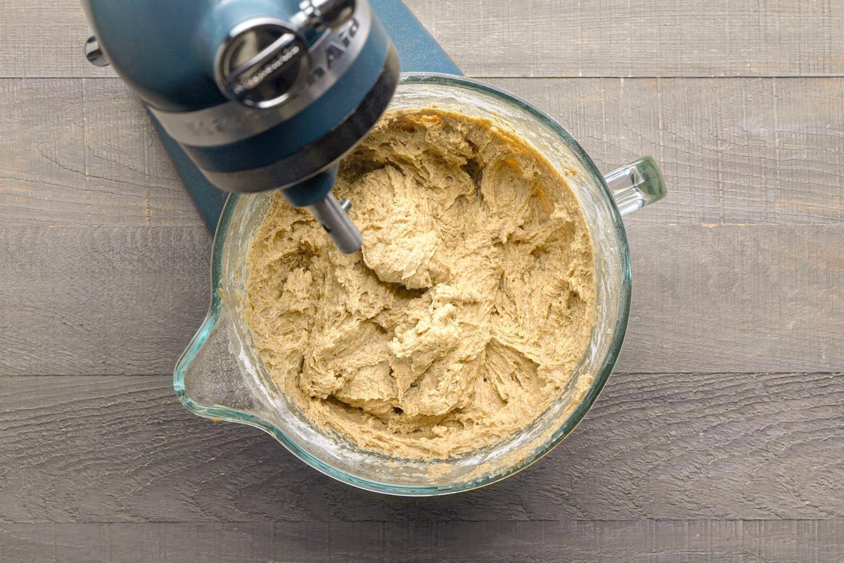 Overhead shot of a stand mixer with a glass bowl mixing a thick, beige dough on a wooden surface;