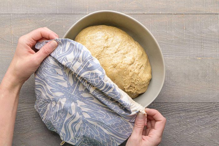 Overhead shot of a person removing a patterned cloth cover from a bowl of risen dough on a light wooden surface;