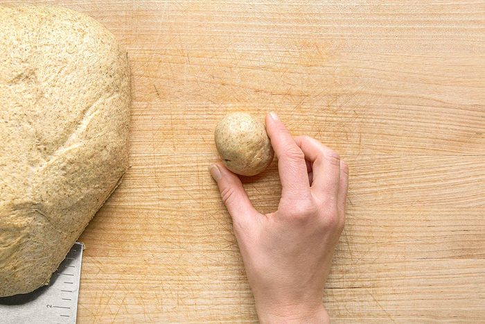 Overhead shot of a hand shaping a small ball of dough on a wooden surface, with a larger piece of dough nearby and a dough scraper partially visible;