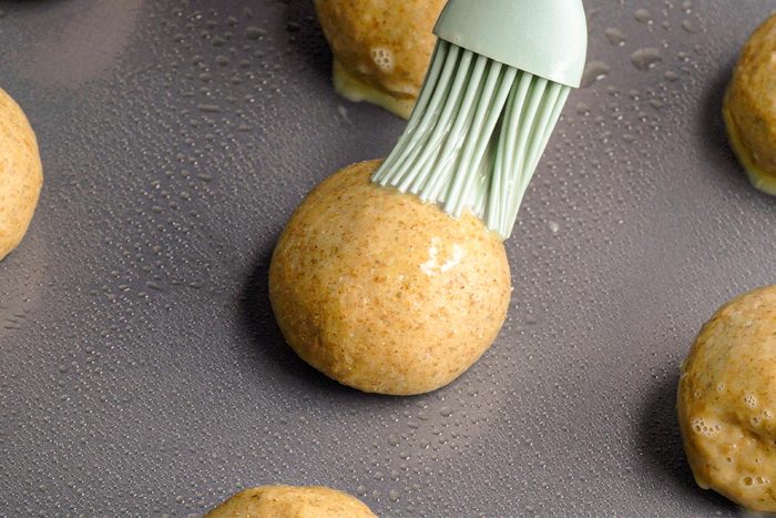Closeup shot of a silicone brush coating a round ball of dough with egg wash on a greased baking sheet, surrounded by other unbaked dough balls;