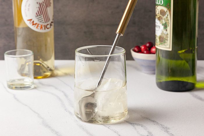Close-up shot of a clear rocks glass filled with Snowy Negroni cocktail is being stirred, with liquor bottles and fresh cranberries visible in the background.