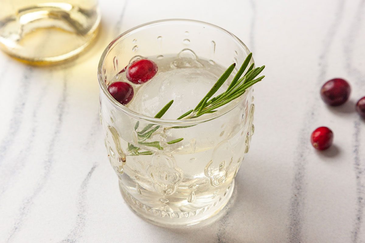 Front-facing shot of a Snowy Negroni served over ice in a rocks glass, garnished with a rosemary sprig and floating cranberries on a marble surface.