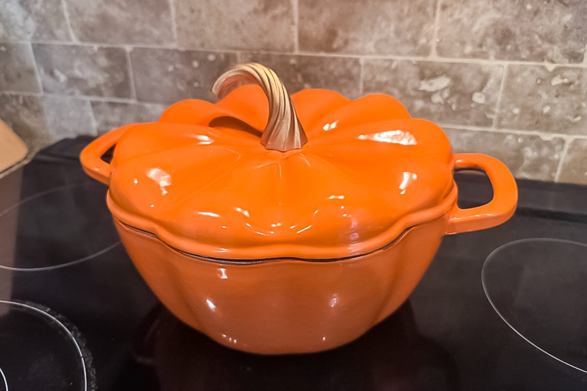 An orange, pumpkin-shaped ceramic pot with a lid and handles sits on a black stovetop in front of a beige tiled backsplash.