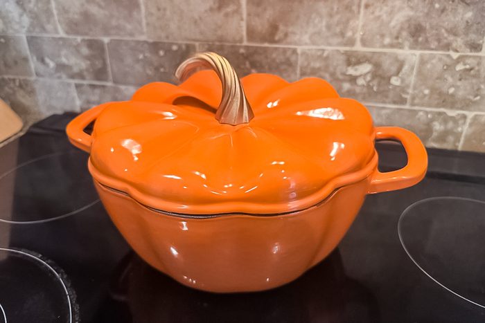 An orange, pumpkin-shaped ceramic pot with a lid and handles sits on a black stovetop in front of a beige tiled backsplash.