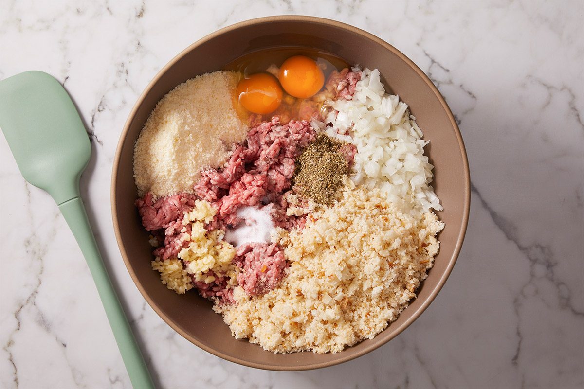 Overhead shot of a bowl sits on a marble surface filled with various ingredients and a pale green spatula rests next to it and Ingredients include ground meat bread crumbs chopped onions eggs grated cheese minced garlic and seasonings