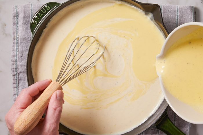 Overhead shot of a hand whisks a creamy yellow sauce in a green skillet as more sauce is poured from a white bowl on a gray-striped cloth and marble surface