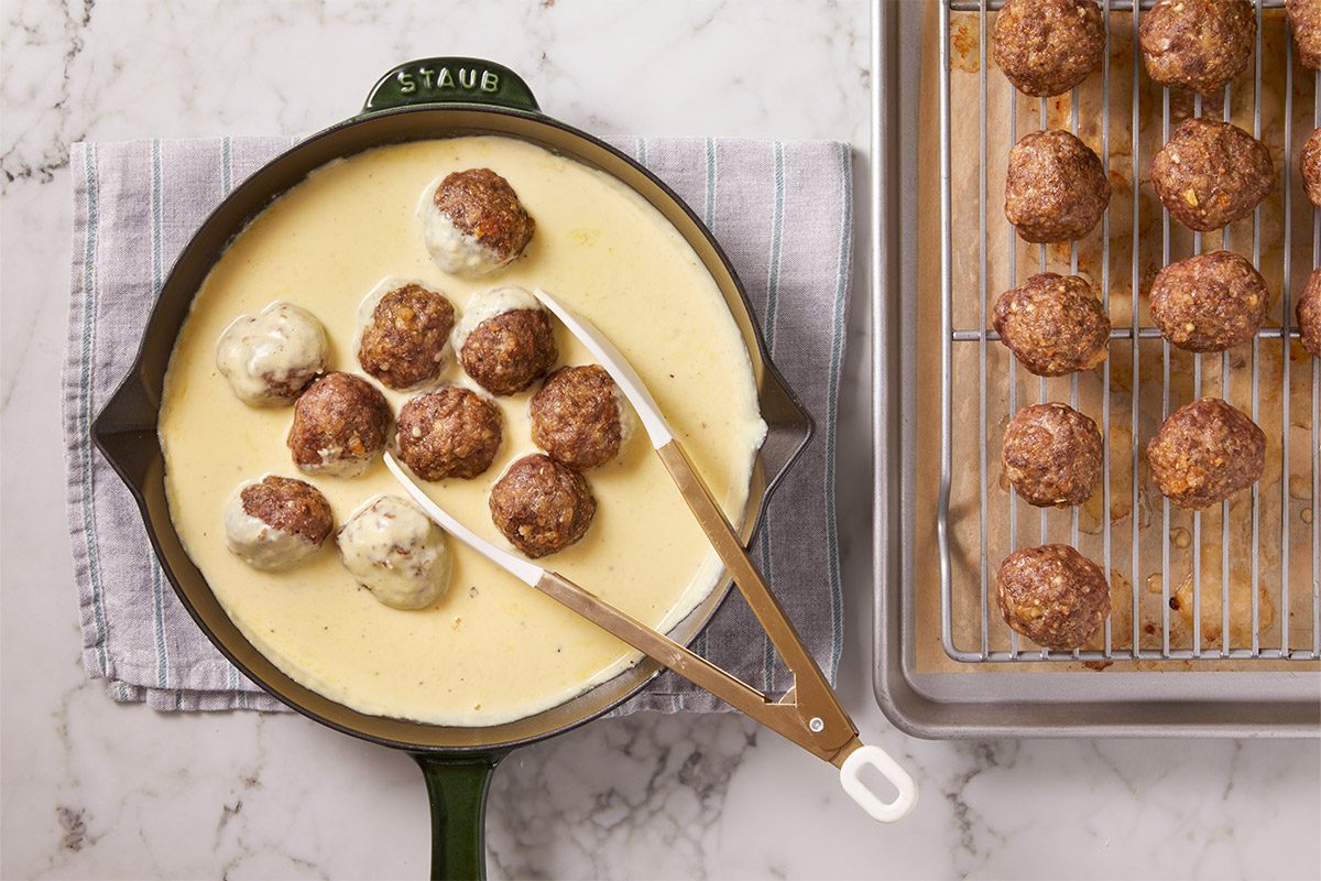 Overhead shot of a skillet with creamy sauce and meatballs rests on a striped towel beside a tray of baked meatballs Tongs lift a meatball in
