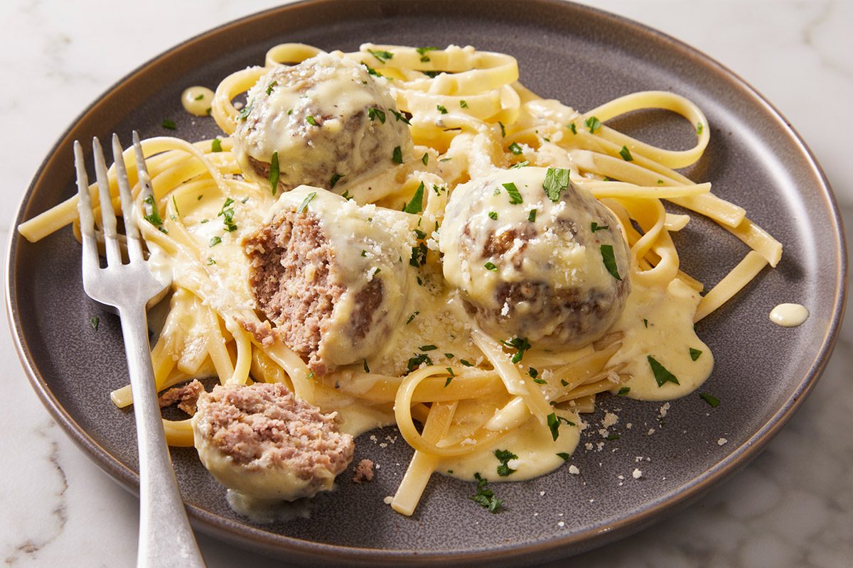 3/4th shot of Alfredo Meatballs with fettuccine pasta and creamy white sauce A cut meatball shows the inside and a fork rests nearby