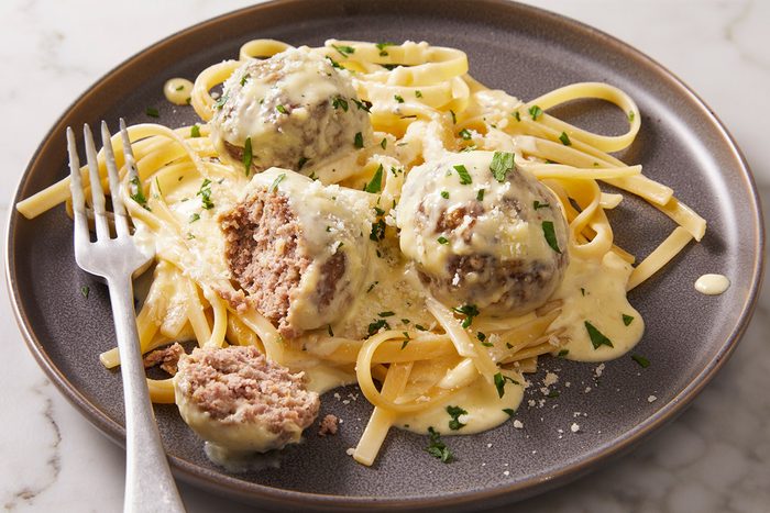3/4th shot of Alfredo Meatballs with fettuccine pasta and creamy white sauce A cut meatball shows the inside and a fork rests nearby